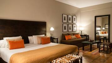 A hotel room with a large bed, a sofa, a small table, a bench, a lamp, and a mirror.