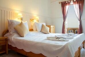 Una cama bien hecha con sábanas blancas y varios almohadones, una mesita de noche de madera con una lámpara, y vistas a un balcón a través de cortinas translúcidas.