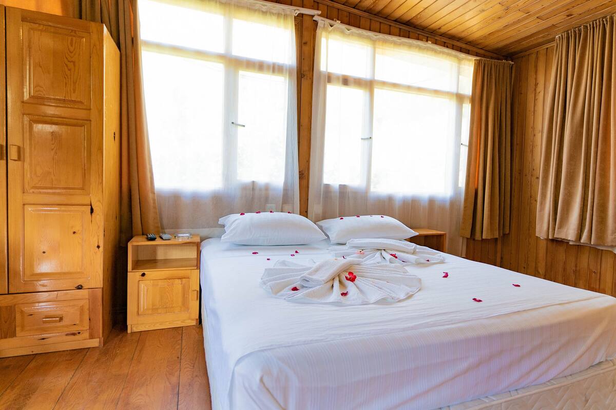 Une chambre d’hôtel avec un plancher en bois, un grand lit avec du linge de lit blanc, deux tables de chevet, une armoire en bois et de grandes fenêtres avec des rideaux.