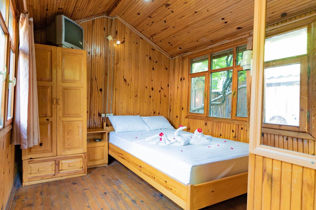 Une chambre dans un chalet en bois, avec un lit, un téléviseur posé sur un meuble et une fenêtre donnant sur la forêt.