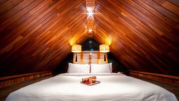 A bedroom with a sloped ceiling, wooden beams, a bed with white linens, two bedside lamps, and a nightstand.