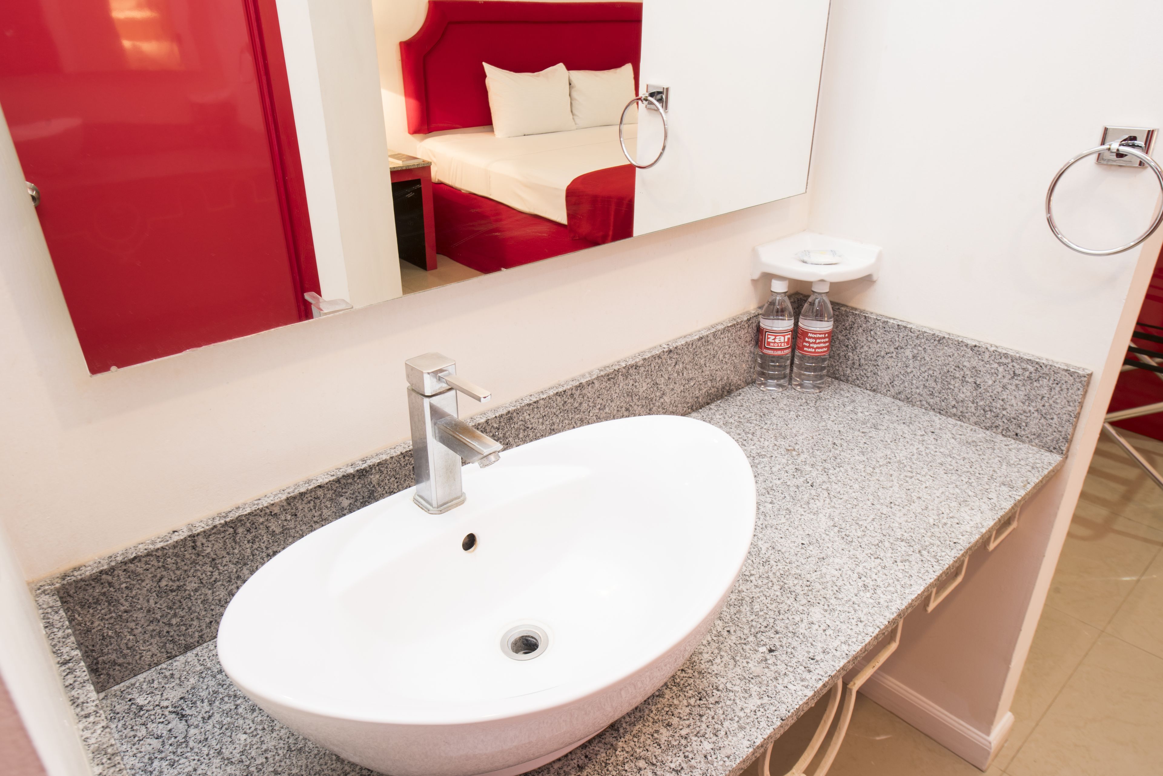Un baño con un lavamanos blanco, un espejo y una cama roja visible en el reflejo.