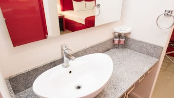 Un baño con un lavamanos blanco, un espejo y una cama roja visible en el reflejo.