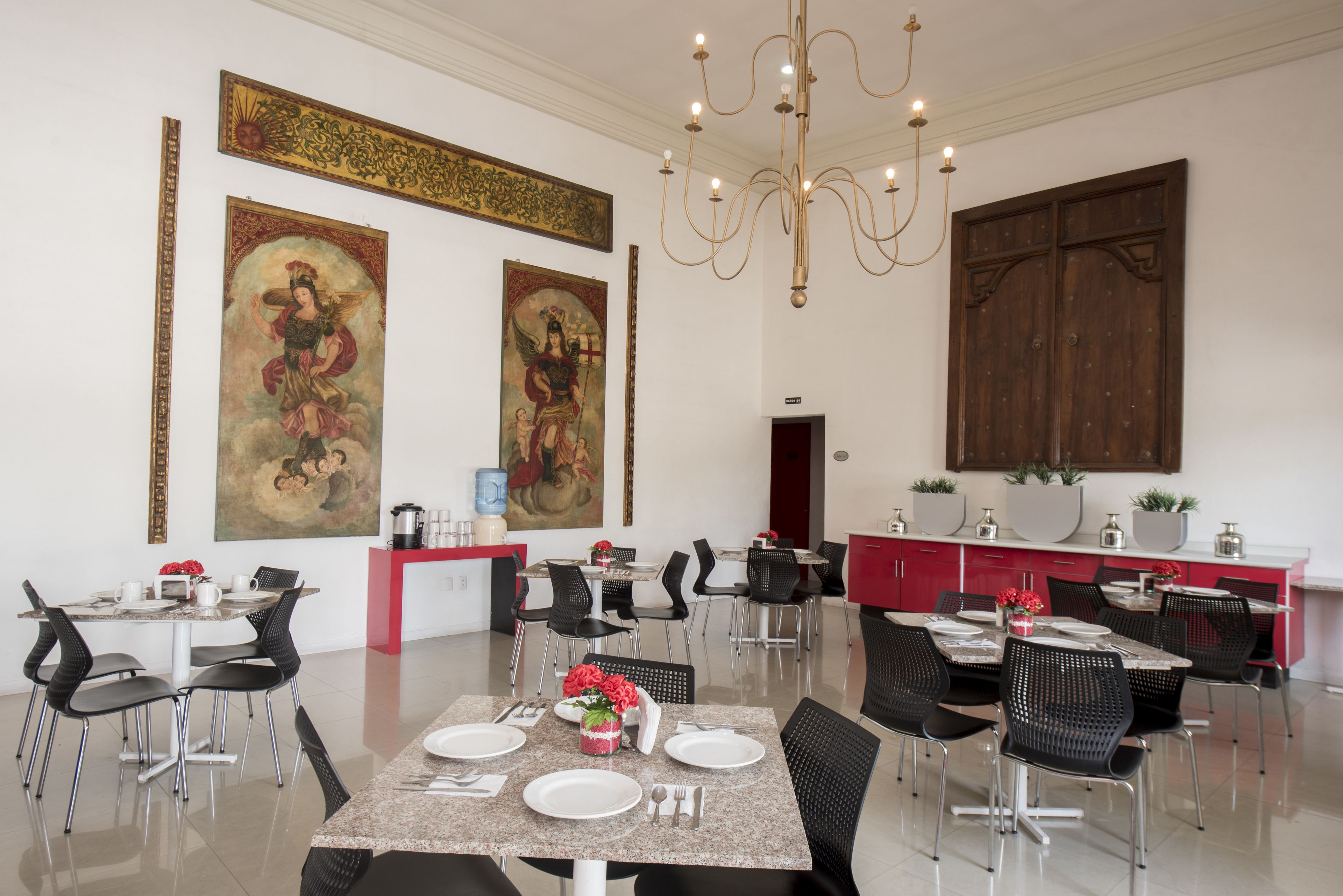 Un comedor con mesas dispuestas para cuatro, sillas negras, una araña y obras de arte en las paredes.