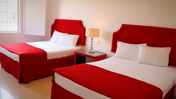 Dos camas rojas con sábanas blancas en una habitación con ventana y una mesita de noche con lámpara.