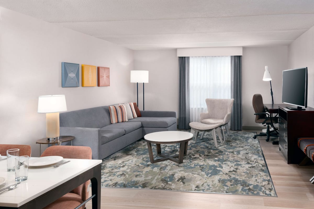 A modern hotel room with a gray sofa, a dining table with chairs, a small round table, a TV, and a large window with curtains.