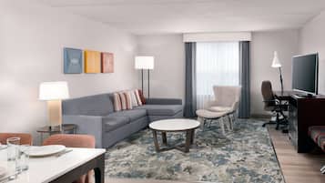 A modern hotel room with a gray sofa, a dining table with chairs, a small round table, a TV, and a large window with curtains.
