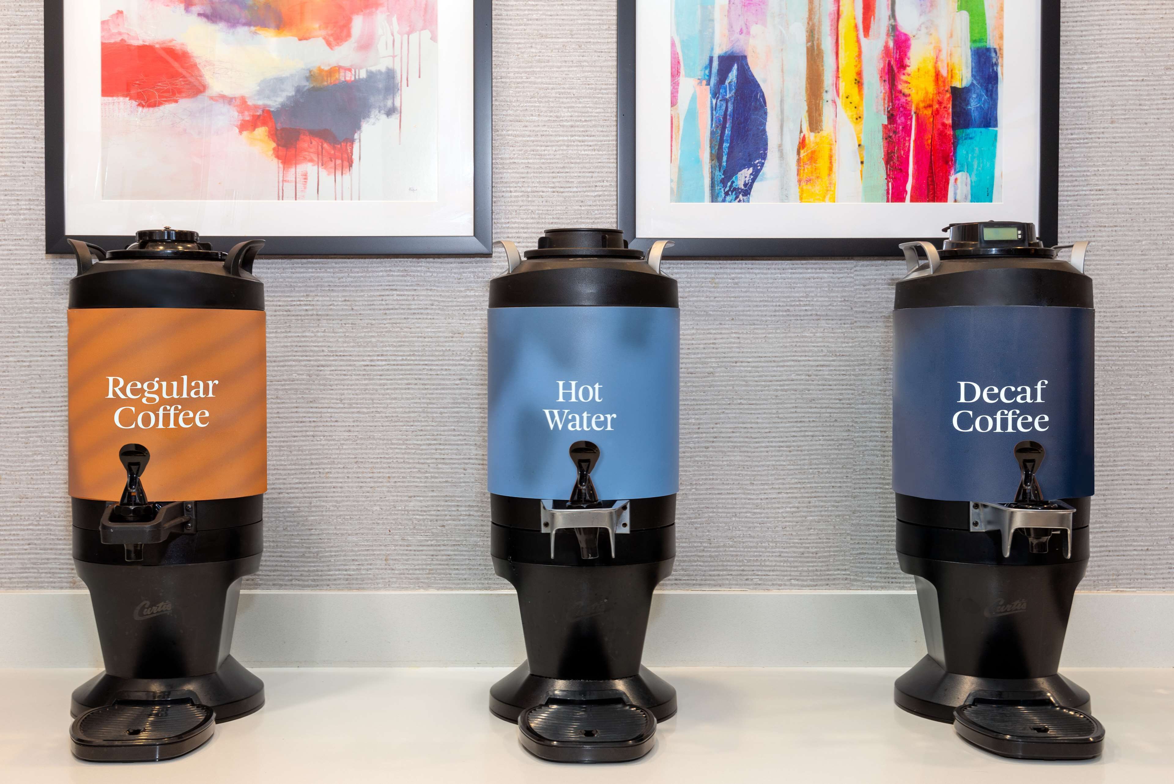 Three coffee dispensers labeled "Regular Coffee," "Hot Water," and "Decaf Coffee" against a wall with abstract art.