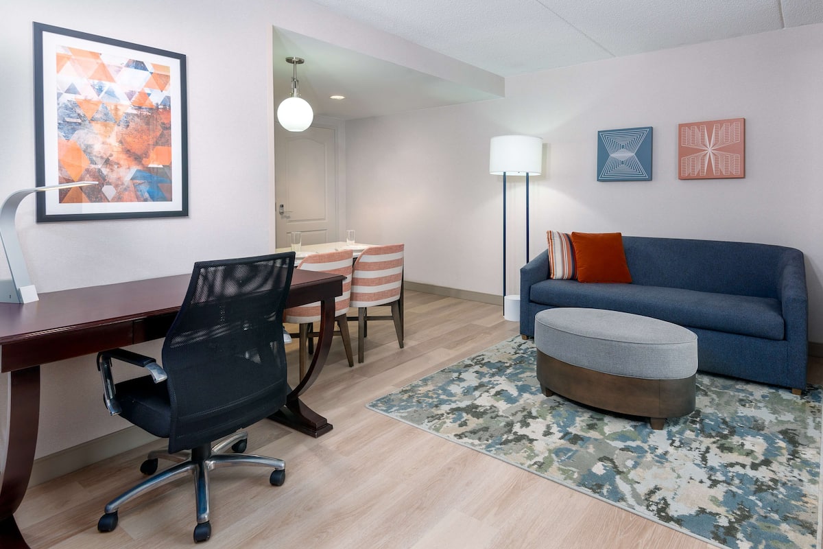 A modern living room with a desk, office chair, sofa, ottoman, and a dining area with chairs and a table.
