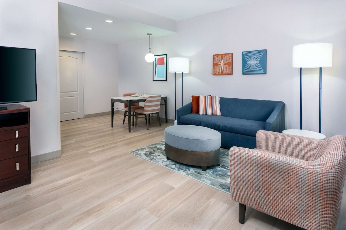 A modern living room with a sofa, ottoman, and armchair, a dining area with chairs, a TV, and artwork on the walls.