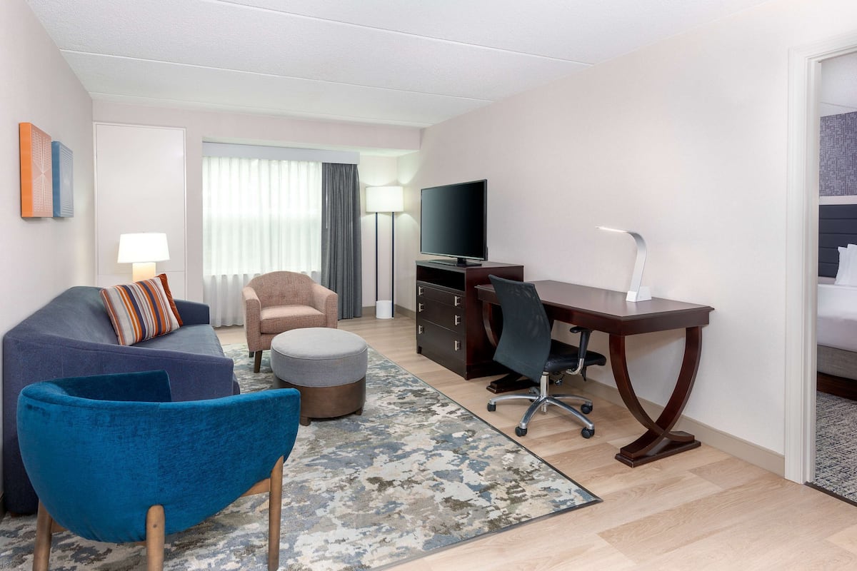 A modern hotel room with a sofa, armchair, ottoman, desk, and television.