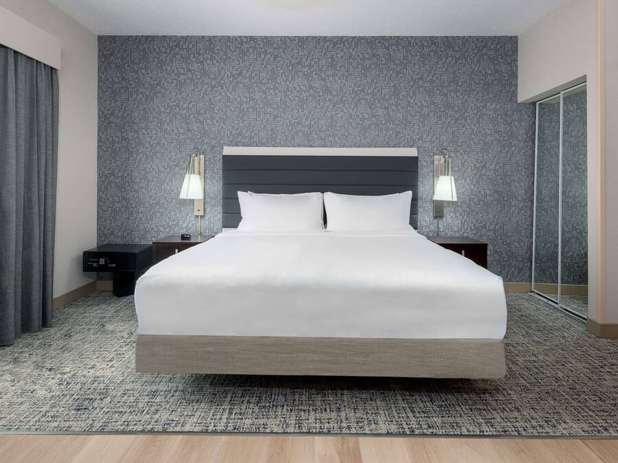 Una cama grande con cabecera oscura, dos mesitas de noche con lámparas, pared con textura y suelo alfombrado.