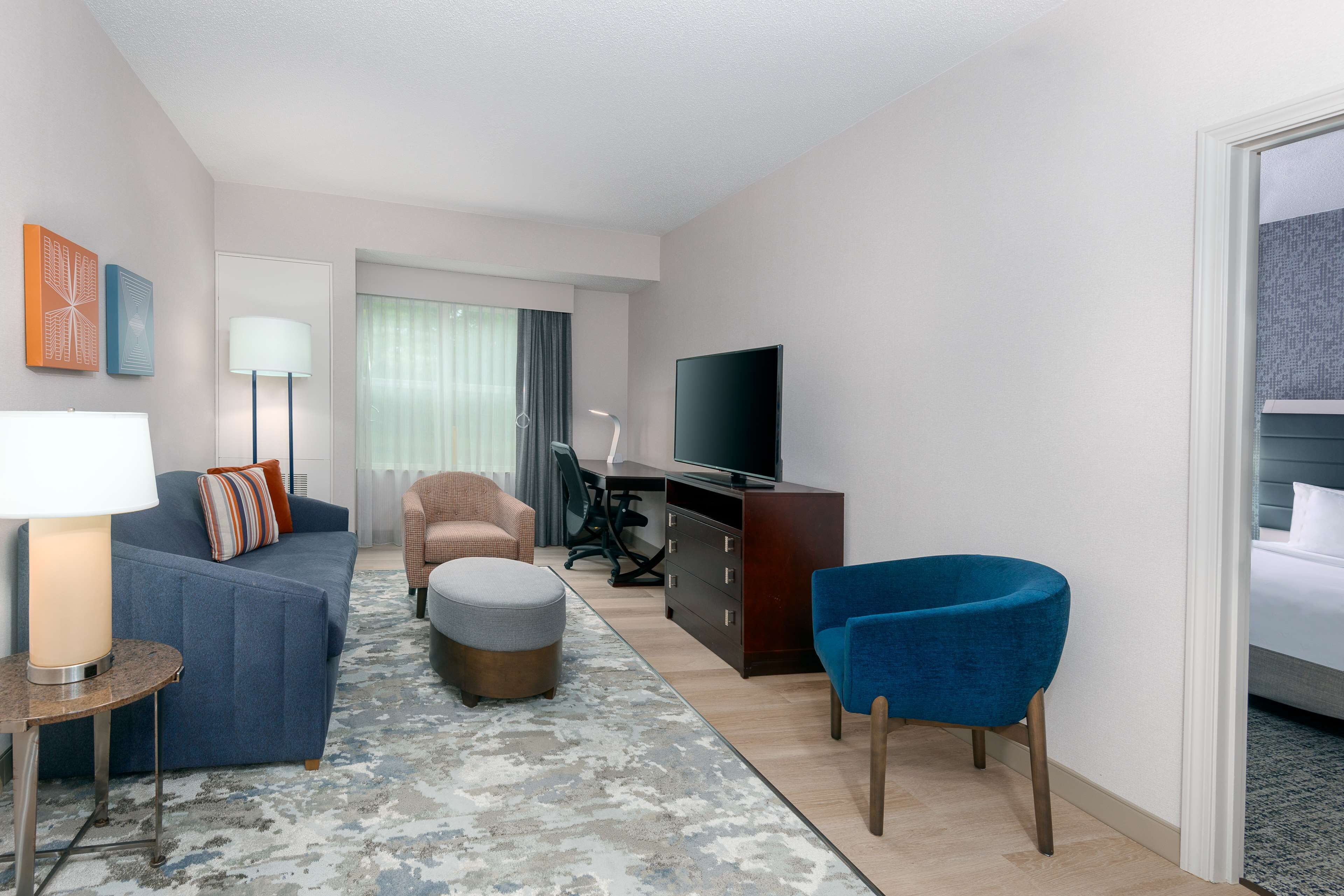 A modern hotel room with a sofa, ottoman, armchair, TV, and a desk with a lamp.