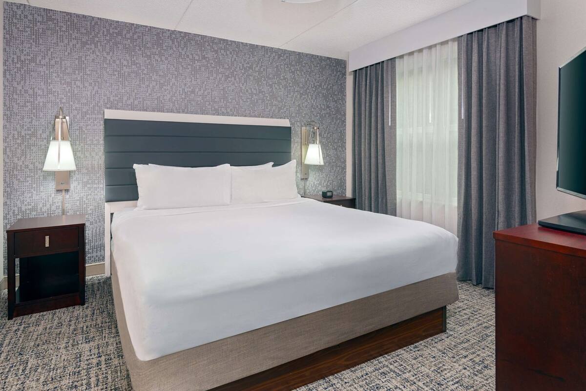 A hotel room with a large bed, two bedside tables, a TV, and a window with curtains.