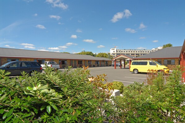 A parking lot with several cars, including a yellow van, in front of a building with a red roof and a white building in the background. - Omahu Motor Lodge (Hastings)