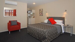 Habitación de hotel con una cama, un sillón rojo, una kitchenette pequeña y una ventana con cortinas.