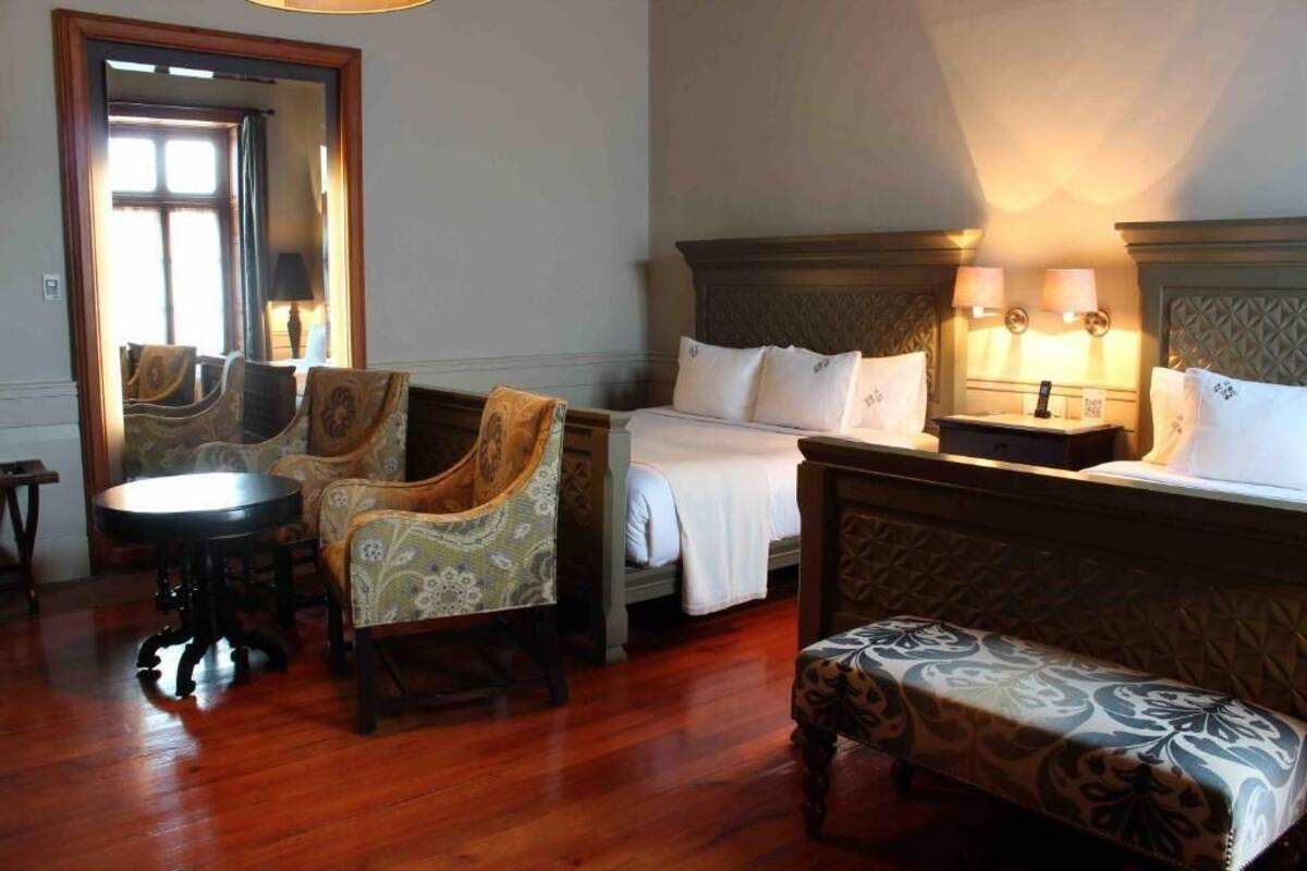 Habitación de hotel con piso de madera, cama con ropa blanca, dos sillones tapizados, una mesita redonda y un banco.