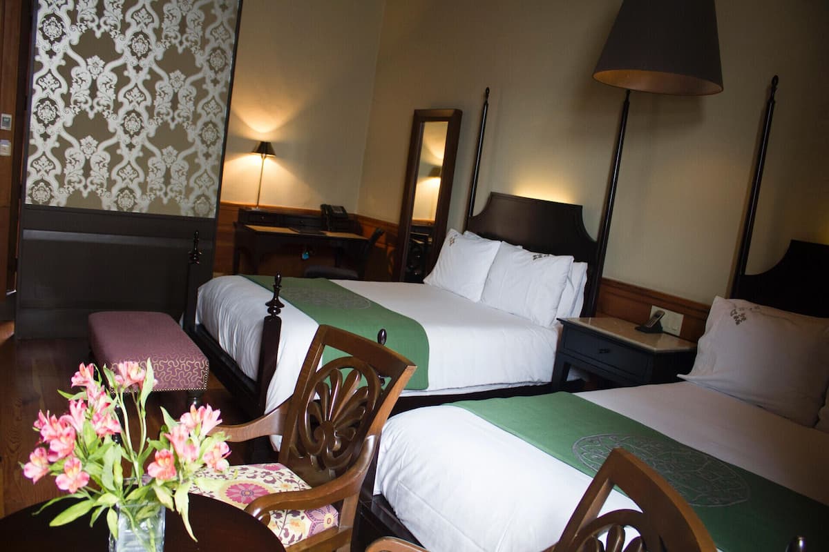 Una habitación de hotel con dos camas, un escritorio, una silla, una lámpara y un florero con flores.