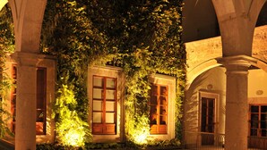 Un patio exterior con un arco de piedra, una pequeña fuente y un edificio bien iluminado con contraventanas de madera.
