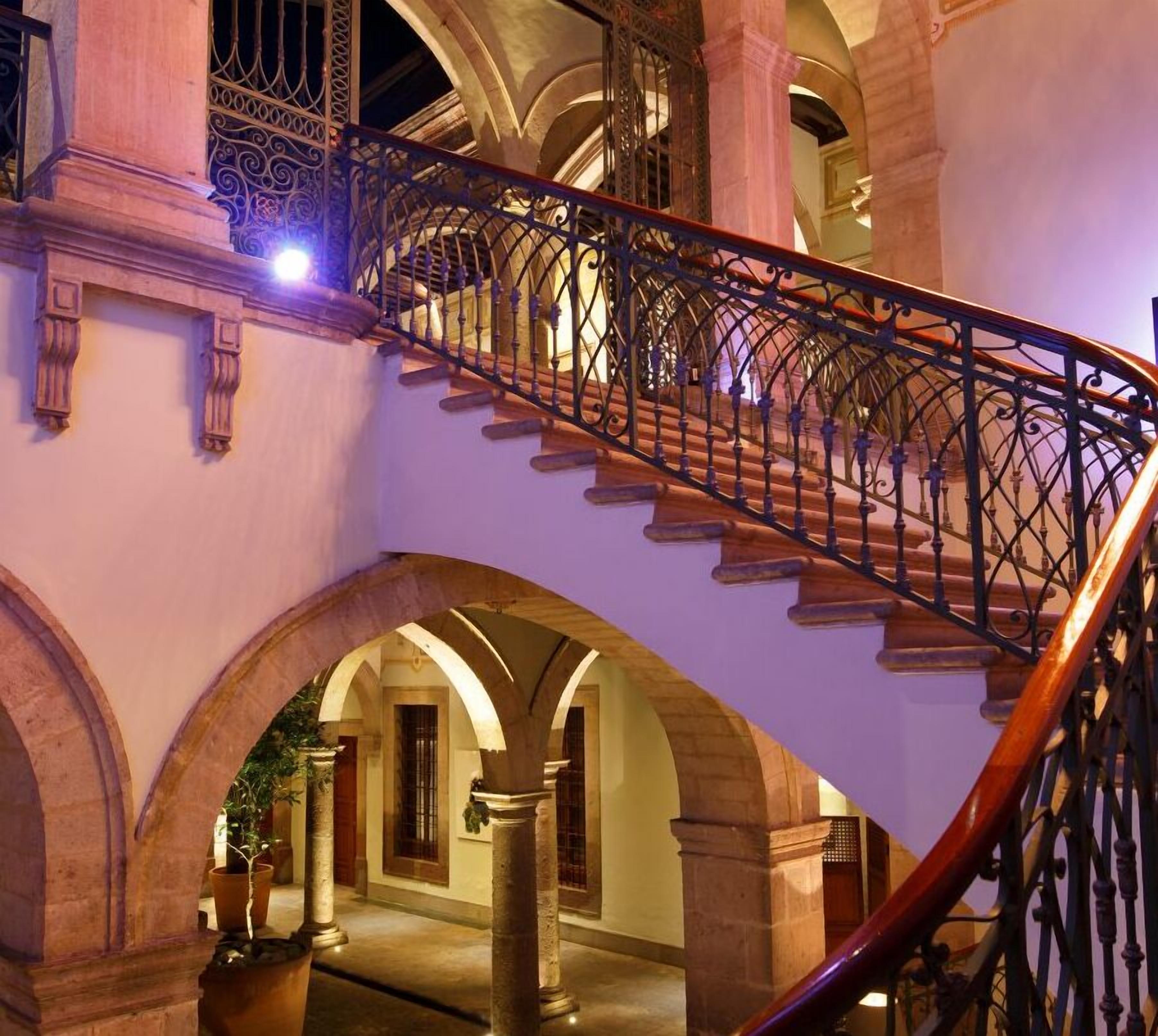 A grand staircase with ornate metal railings and stone balustrades, leading to an upper level with arched doorways and columns.