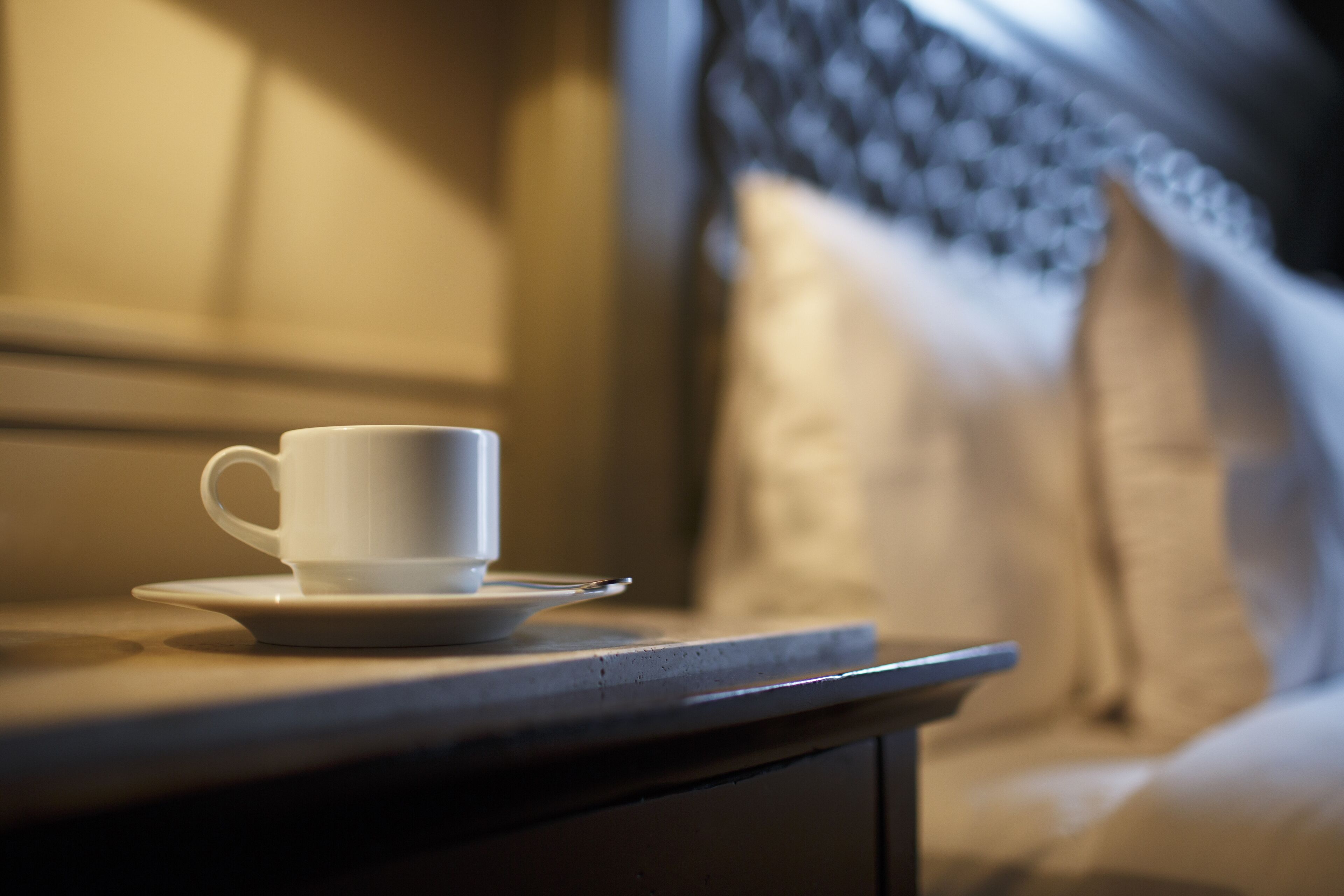 Une tasse et une soucoupe blanches sont posées sur une table de chevet dans une pièce faiblement éclairée, avec une tête de lit à motifs et des coussins.