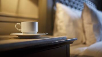 Une tasse et une soucoupe blanches sont posées sur une table de chevet dans une pièce faiblement éclairée, avec une tête de lit à motifs et des coussins.