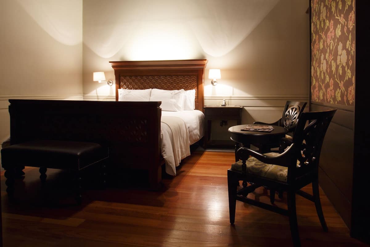 Una habitación de hotel con una cama de madera, una mesita redonda pequeña, una silla y un banco.