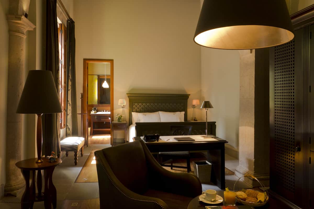Habitación de hotel con cama, escritorio, silla, lámpara y mesa con una taza y una cesta de pan.
