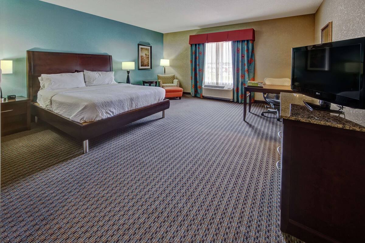 A hotel room with a large bed, a desk with a TV, a chair, and a window with curtains.