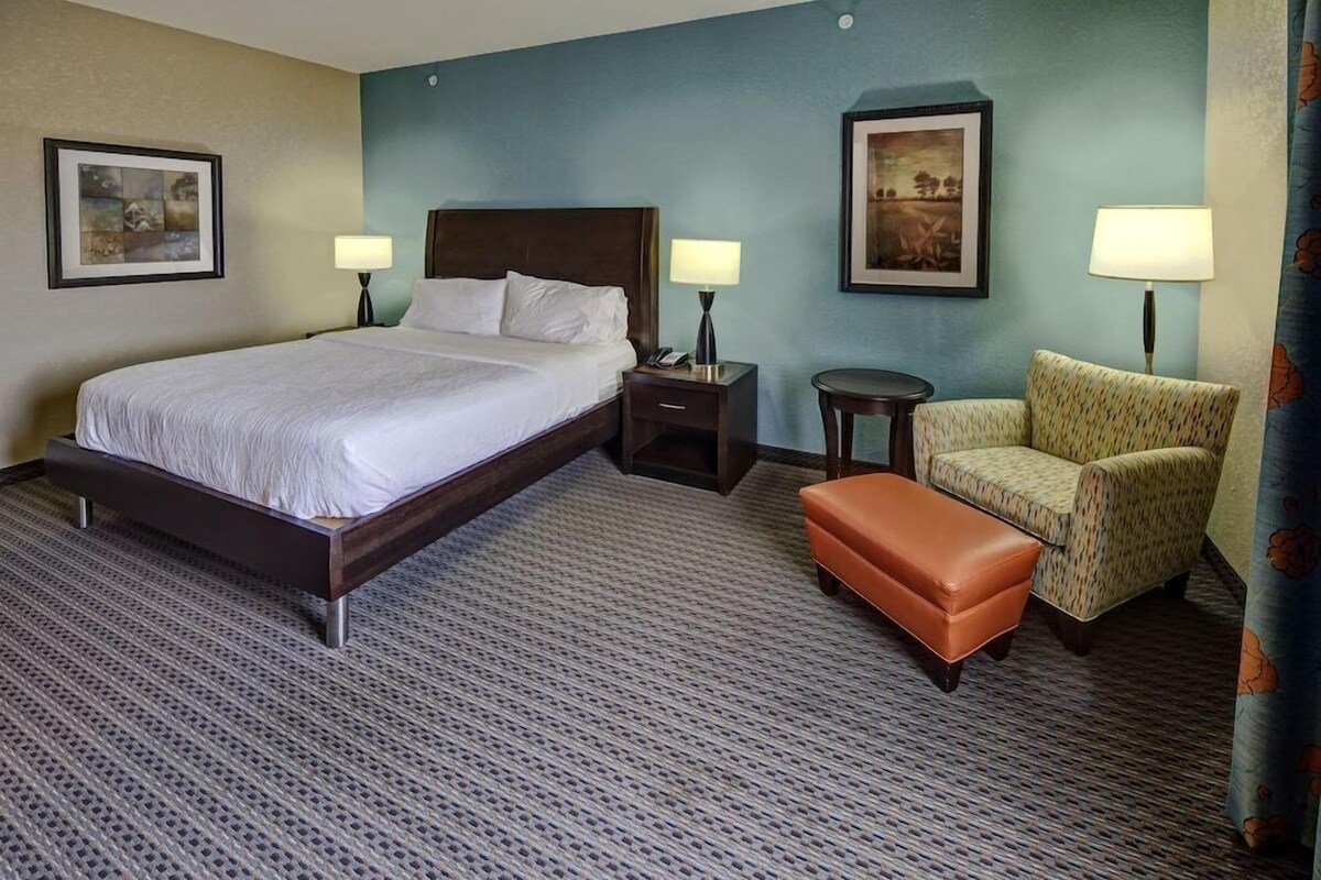 A hotel room with a large bed, two bedside tables with lamps, a chair, an ottoman, and two framed pictures on the walls.