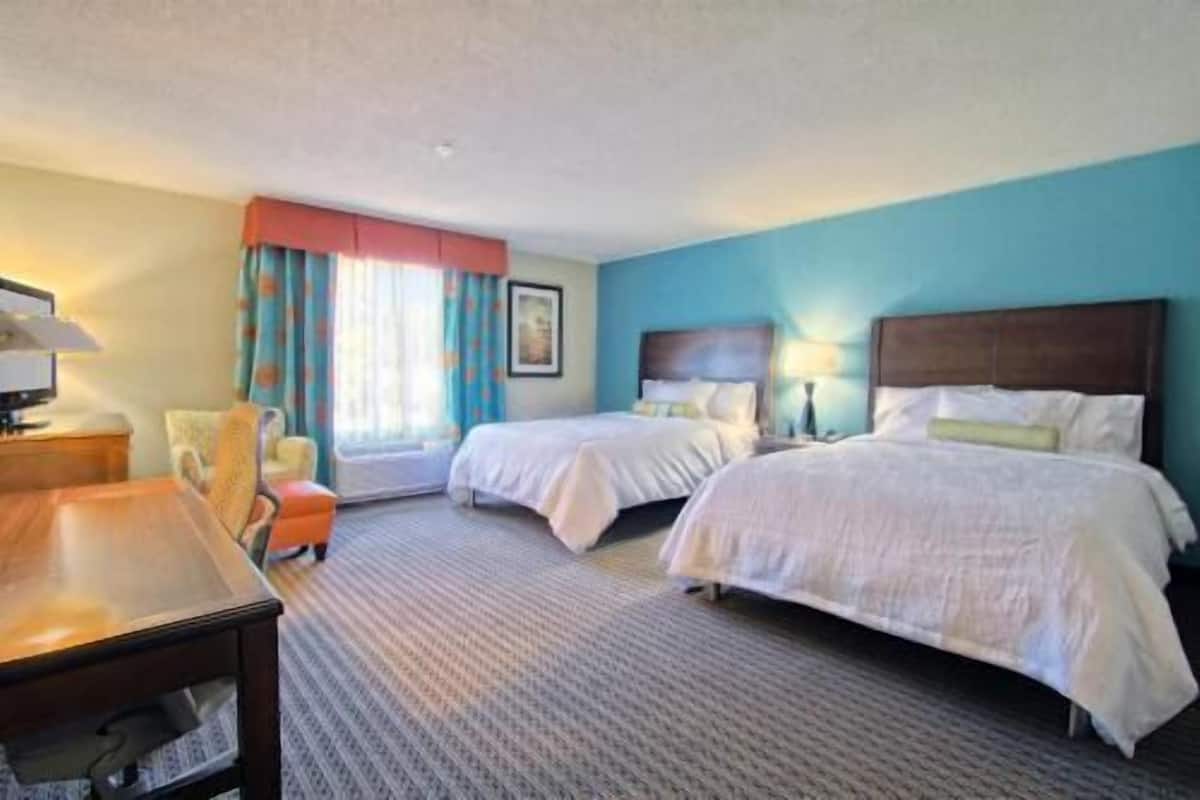 A hotel room with two beds, a desk, a chair, and a window with curtains.