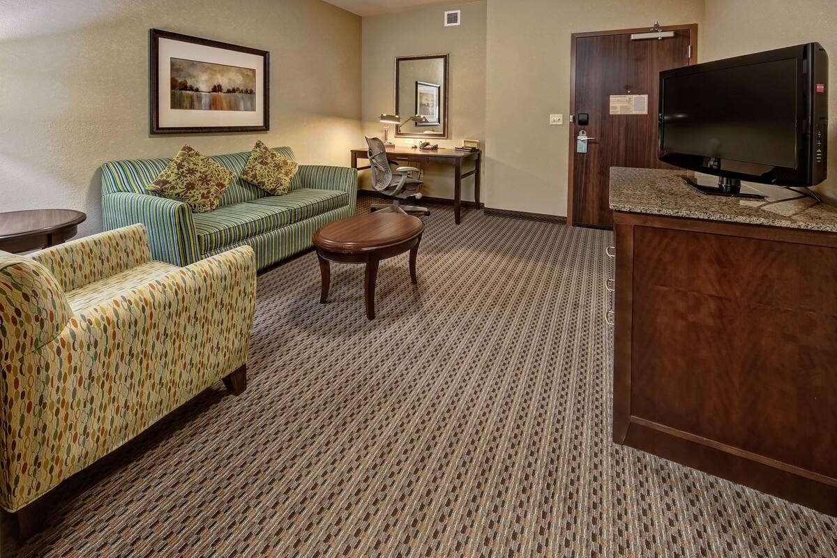 A hotel room with a green-striped sofa, a patterned armchair, a small wooden table, a desk with a chair, a television, and a door with a keycard lock.