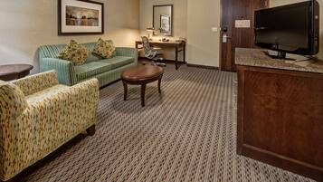 A hotel room with a green-striped sofa, a patterned armchair, a small wooden table, a desk with a chair, a television, and a door with a keycard lock.