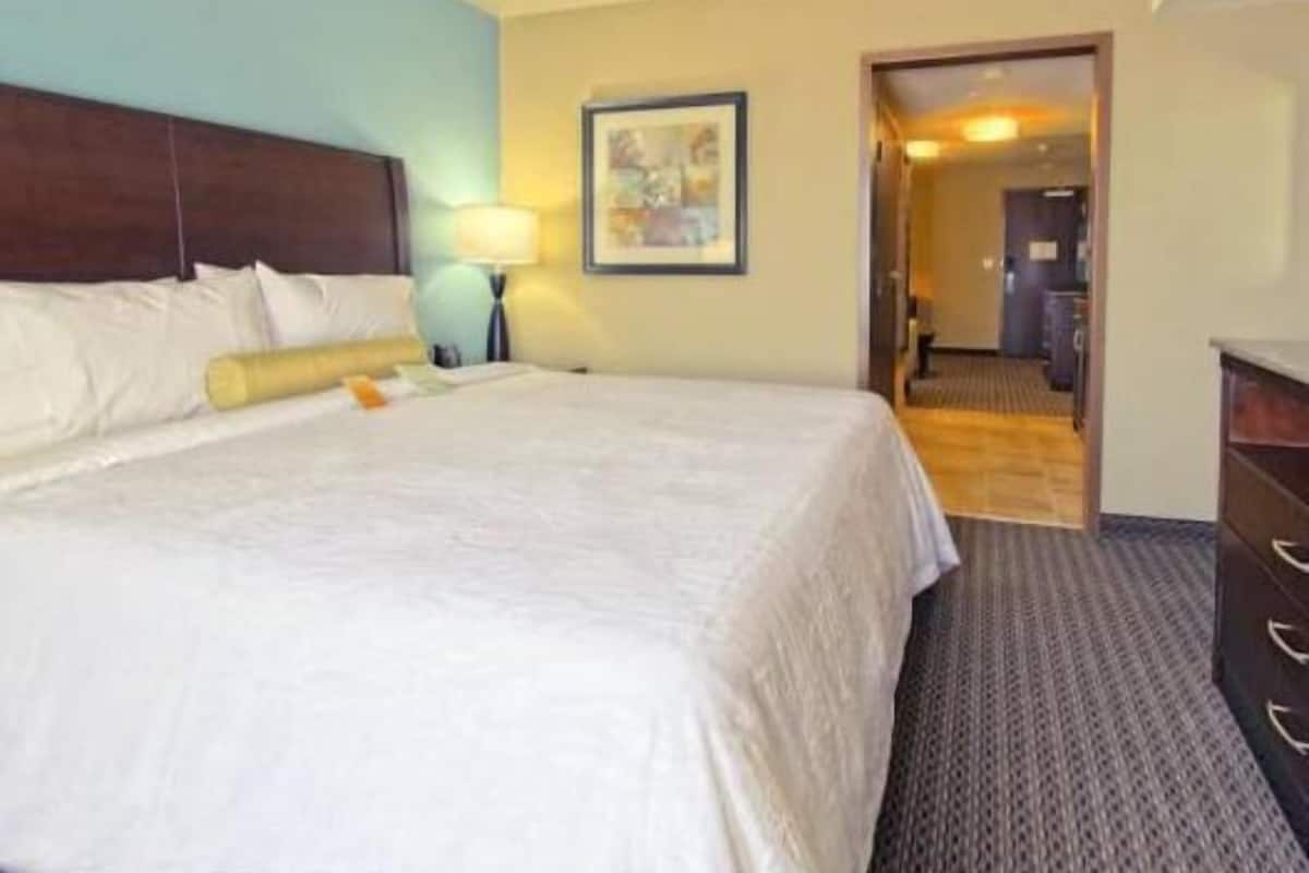 A hotel room with a large bed, a nightstand, a lamp, a framed picture on the wall, and a door leading to another room.