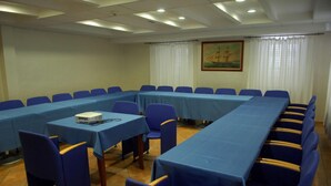 Een vergaderzaal met lange tafels, blauwe stoelen, een beamer en een schilderij aan de muur.