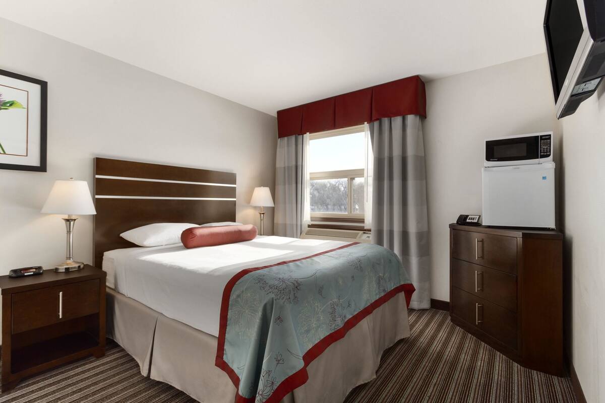 Ett hotellrum med en säng, två sängbord, ett skrivbord, en mikrovågsugn och ett fönster med gardiner.