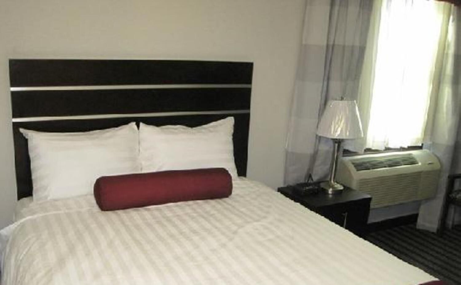 A hotel room with a bed, a red pillow, a nightstand with a lamp, and an air conditioning unit.
