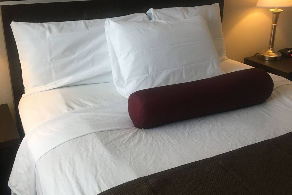 Ett hotellrum med en säng, vita sängkläder, en röd kudde, ett nattduksbord med en lampa och en mörk sänggavel.