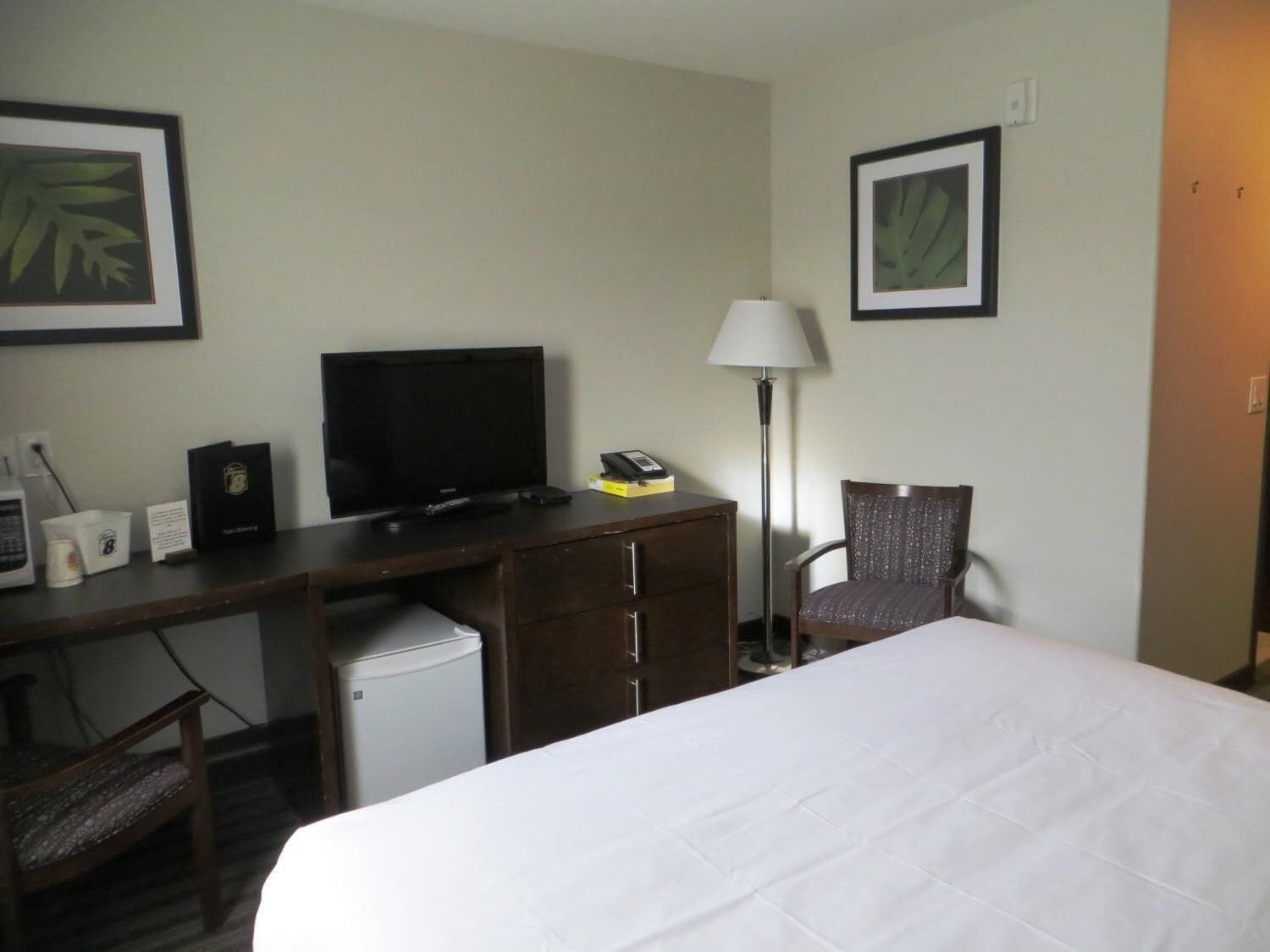 A hotel room with a bed, a desk with a television, a chair, a lamp, and a small refrigerator.