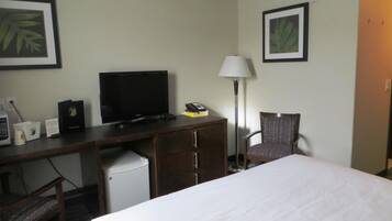 A hotel room with a bed, a desk with a television, a chair, a lamp, and a small refrigerator.