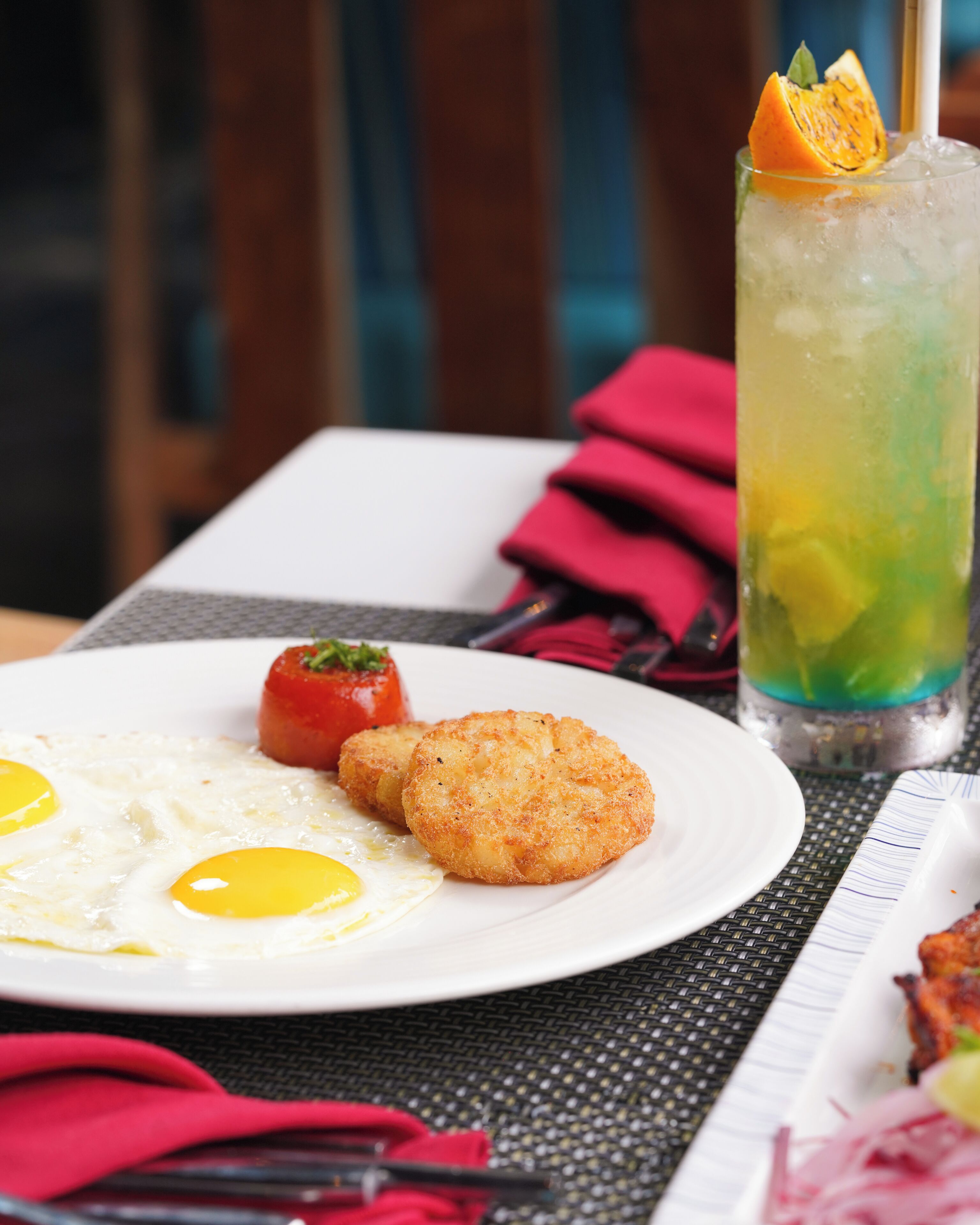 A plate with fried eggs, a tomato, and a breaded item, alongside a tall drink with a fruit garnish.
