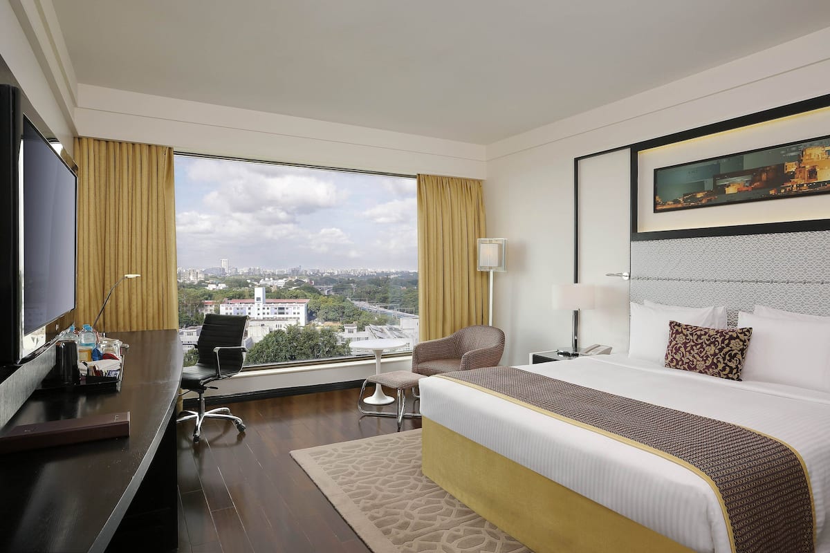A hotel room with a large bed, a desk with a computer, a chair, a TV, and a window with a city view.