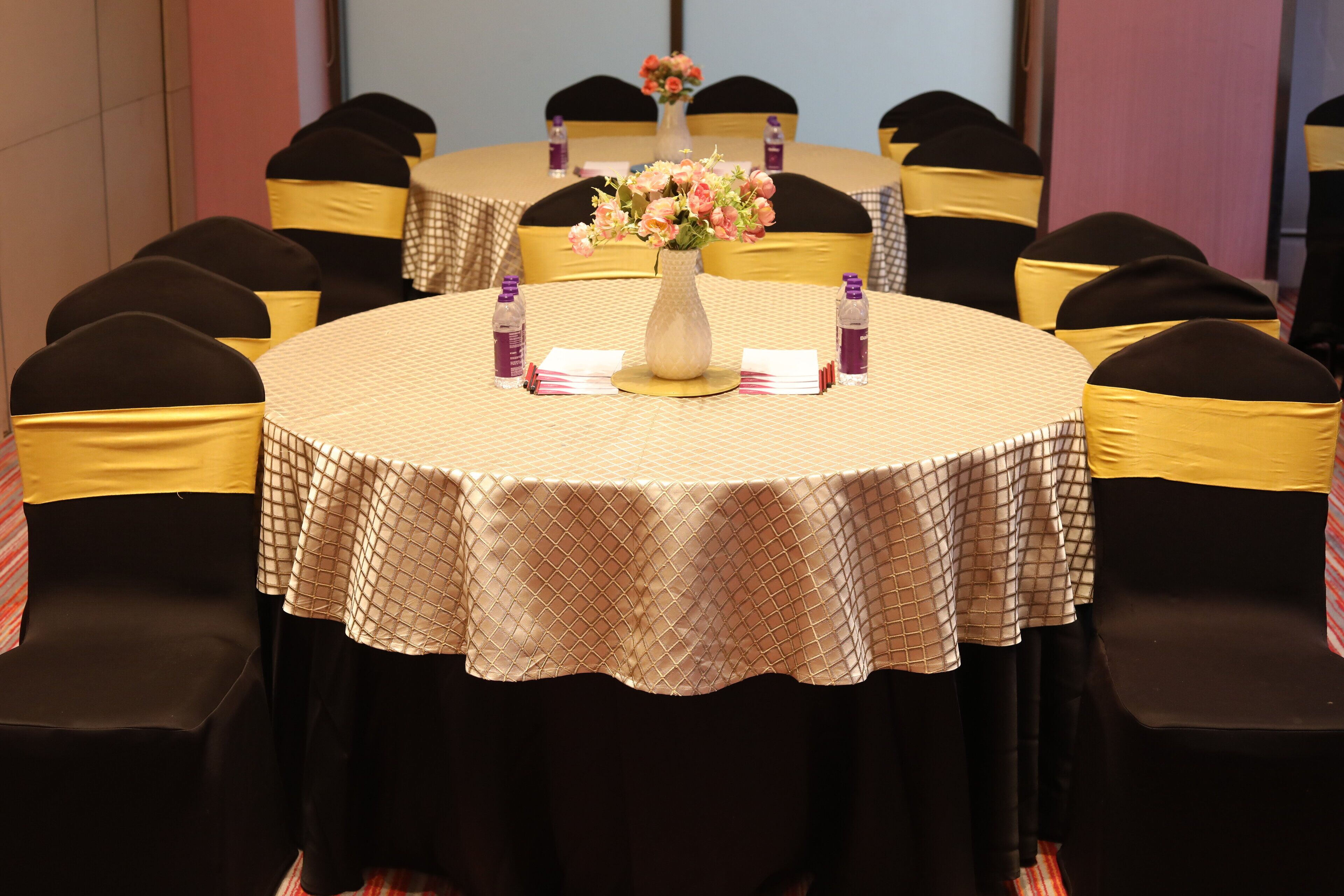 Un salón de conferencias con mesas redondas, sillas negras con cintas amarillas y un jarrón con flores sobre las mesas.