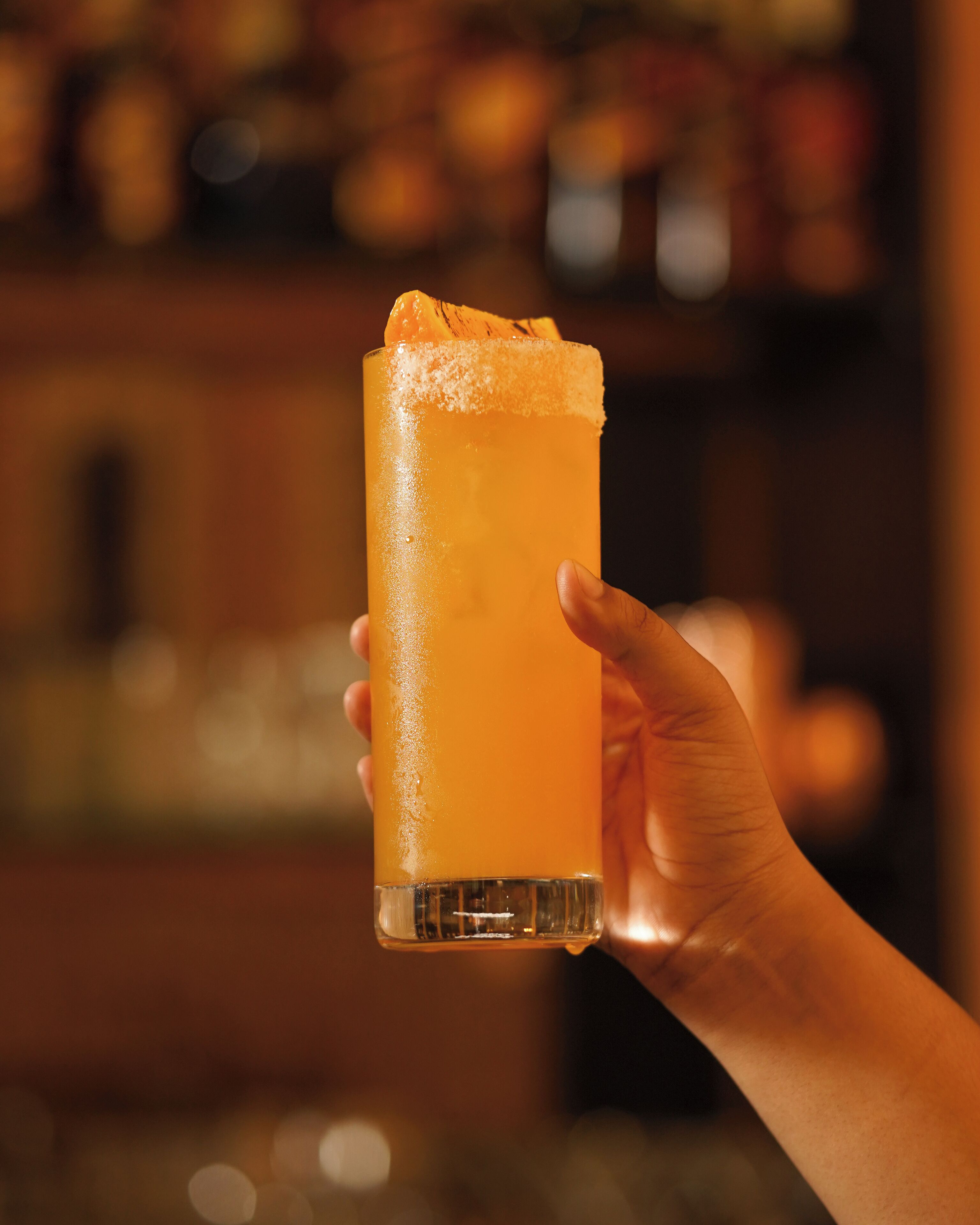Een hand houdt een hoog glas met een schuimend drankje, waarschijnlijk een cocktail, met een onscherpe achtergrond die een bar of restaurant doet vermoeden.