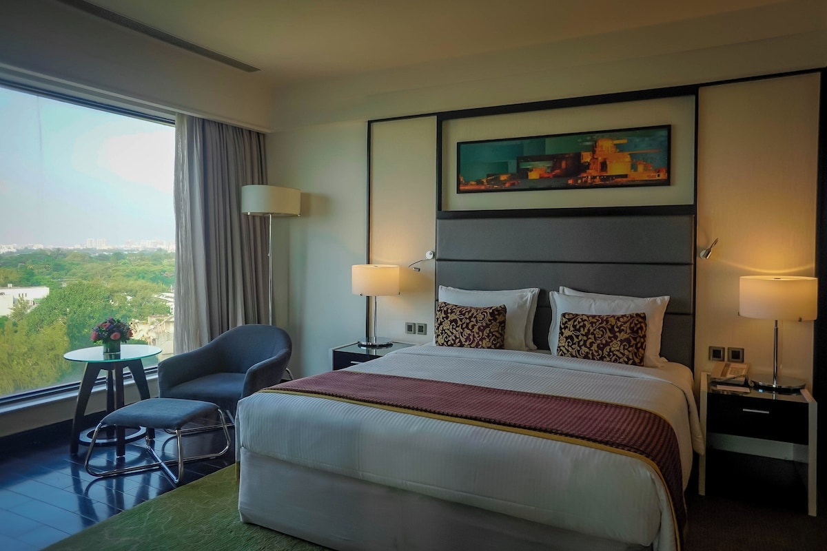 Une chambre d’hôtel avec un grand lit, deux tables de chevet, une chaise, une petite table sur laquelle est posé un vase, et une grande fenêtre donnant sur la ville.