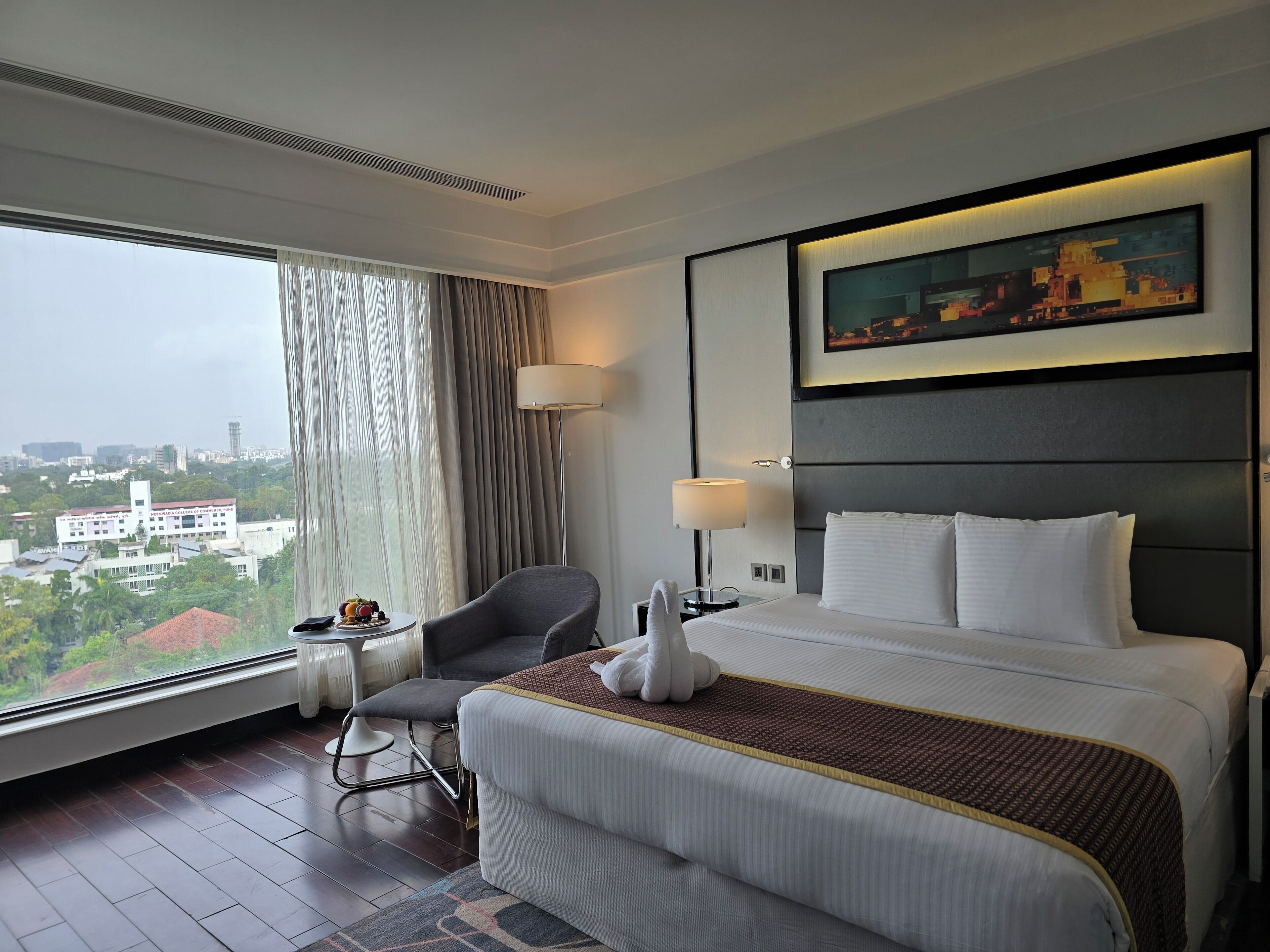 A hotel room with a large bed, a chair, a desk with a lamp, and a view of a cityscape through the window.