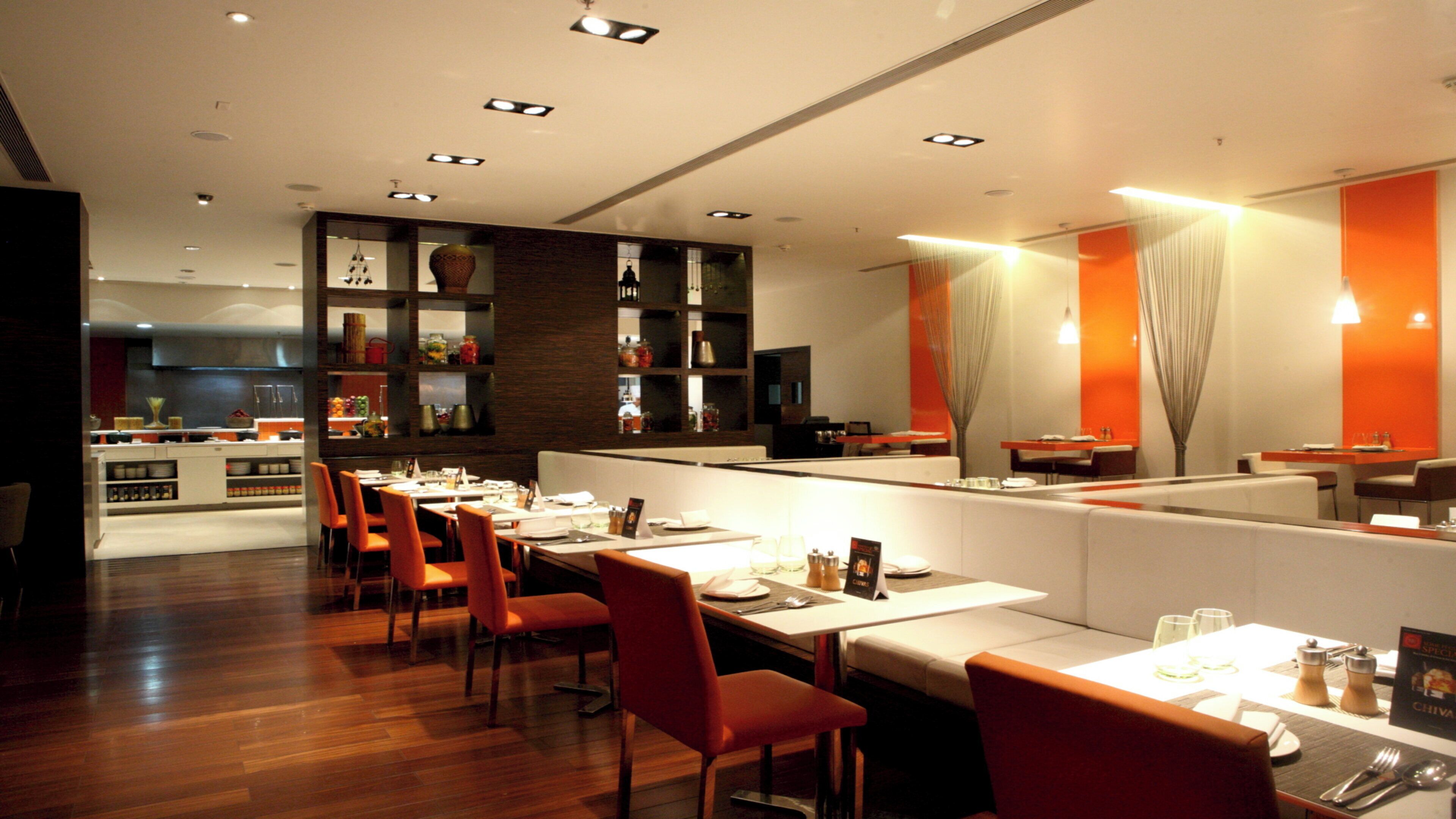 A modern restaurant interior with wooden flooring, orange chairs, and a central dining area with white tables.