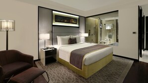 Une chambre d’hôtel avec un grand lit, une table de chevet, une chaise, un bureau, une lampe, un miroir et une salle de bain visible à travers une porte ouverte.