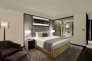 Quarto de hotel com uma cama grande, mesa de cabeceira, cadeira, escrivaninha, abajur, espelho e banheiro visível através de uma porta aberta.
