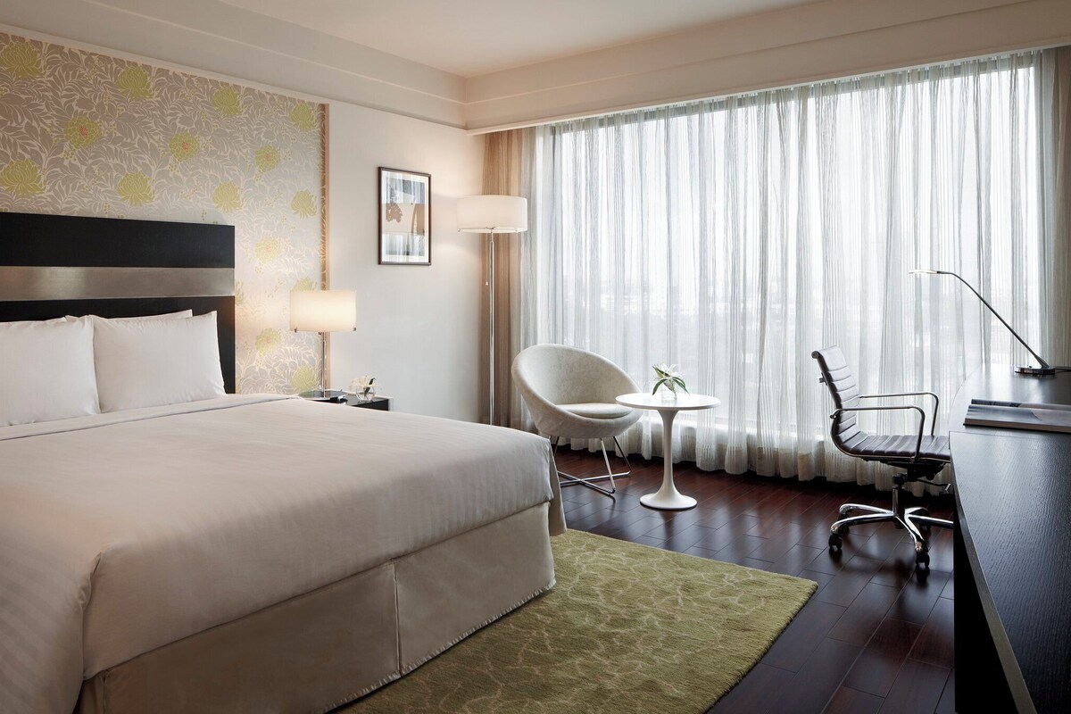 Une chambre d’hôtel avec un grand lit, un bureau, une chaise, une lampe et une vue sur l’extérieur à travers des rideaux transparents.